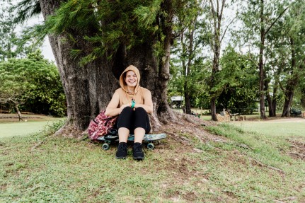 Young person smiling whilst sitting in front of a tree with a skateboard